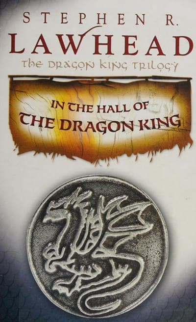 A pale grey-blue field with 'STEPHEN R. LAWHEAD' in large dark red serif capitals at the top, 'THE DRAGON KING TRILOGY' in smaller red serif beneath it, a torn parchment-scroll banner in gold and brown carrying the title 'IN THE HALL OF THE DRAGON KING' in dark red serif, and below it a round silver medallion embossed with a coiled winged dragon.