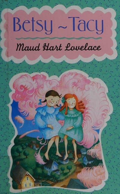 A mint-green paperback jacket with a pink scalloped panel holding the title 'Betsy ~ Tacy' in blue script and the author's name in black script beneath it, over an illustration of two girls in vintage dresses standing amid pink cloud-like puffs above a small hillside town.