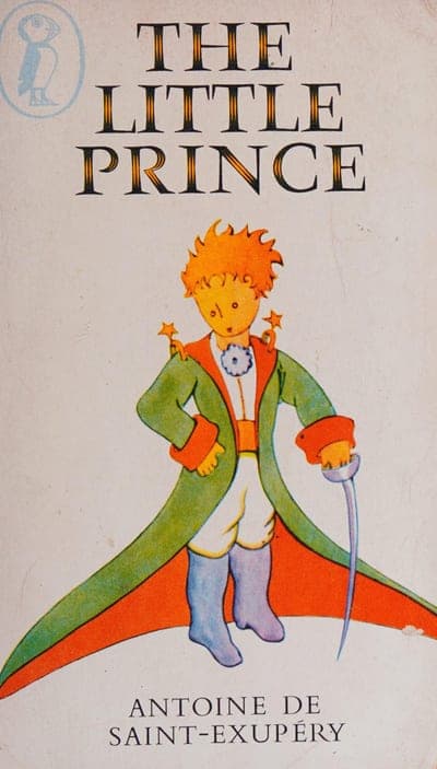 A cream Puffin paperback with the round blue Puffin bird logo at top left, 'THE LITTLE PRINCE' in tall black serif capitals across the top, a full-color illustration of the Little Prince in his green cape and red-lined cloak standing on his tiny planet with a sword at his side, and 'ANTOINE DE SAINT-EXUPÉRY' in black capitals at the bottom.