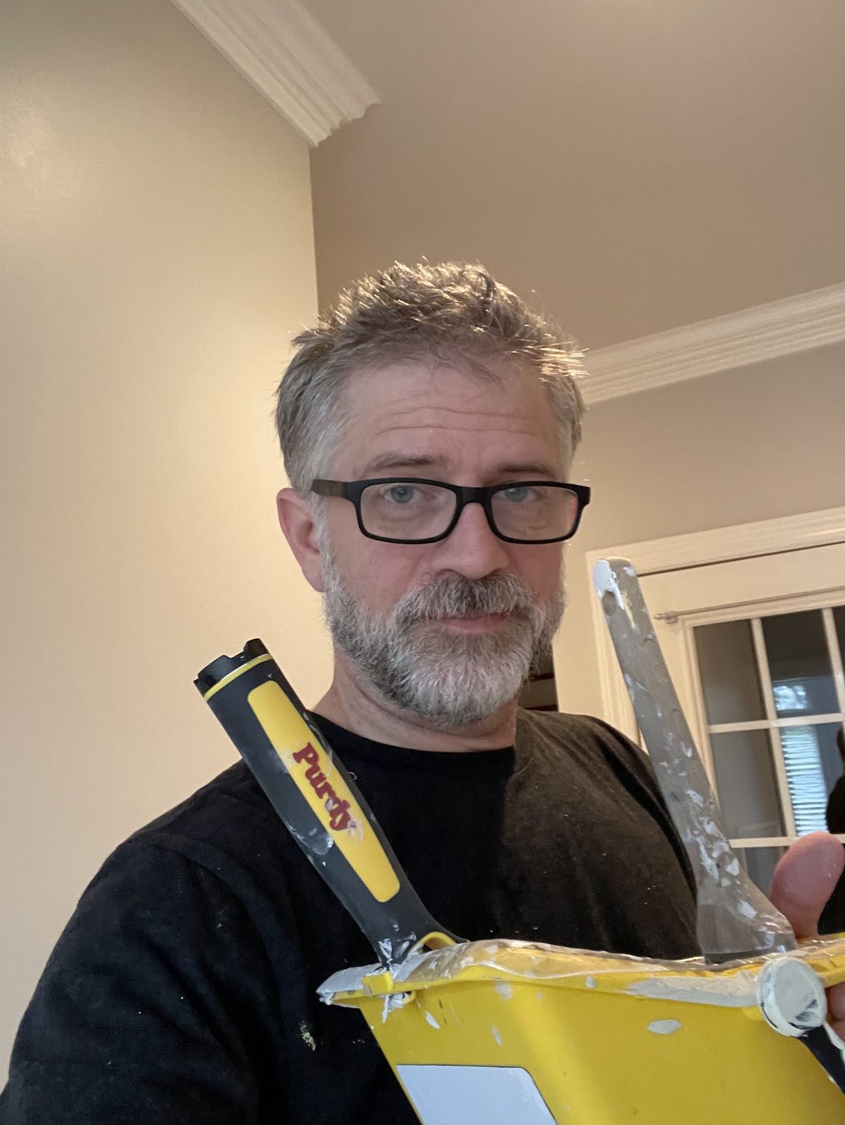 David Clark in glasses, holding a yellow trim bucket and a Purdy brush, with a freshly painted room and crown molding behind him