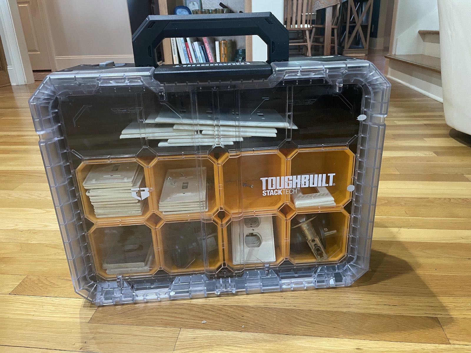 A clear-lidded organizer holding switch plates, outlet covers and door hardware sorted into bins