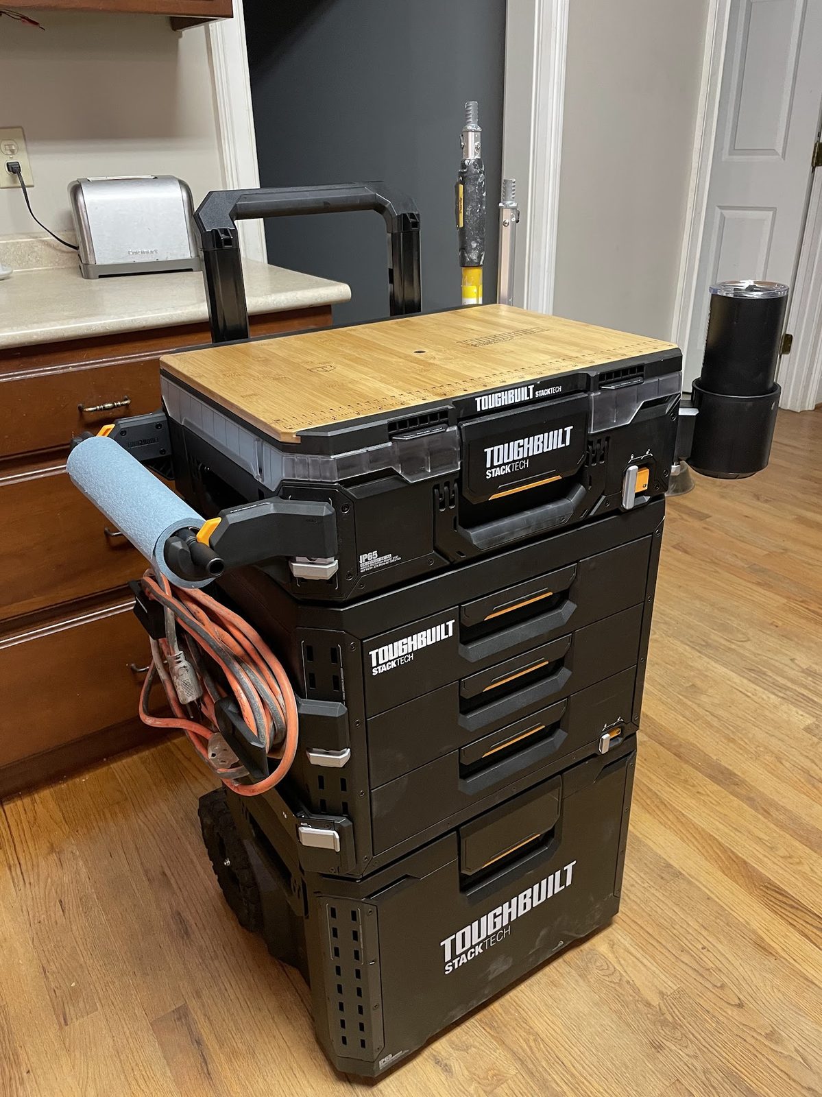 A rolling black tool cart with a wooden top, an extension cord and a paint roller hung on the side, standing on a hardwood floor